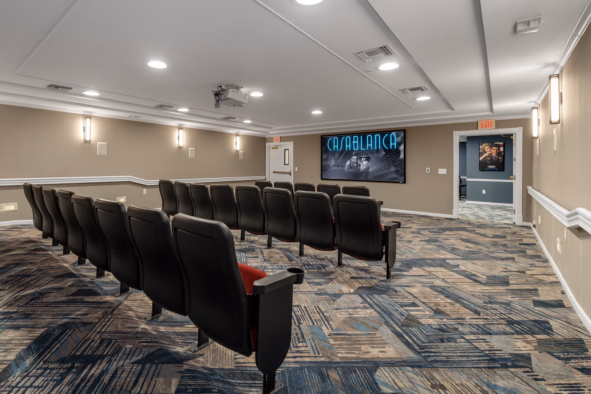 A small movie theater room with two rows of black cushioned seats facing a large screen displaying the movie title 'Casablanca'. The room has patterned carpet flooring, beige walls with white trim, and ceiling lights. There is a projector mounted on the ceiling and an exit door at the back of the room.