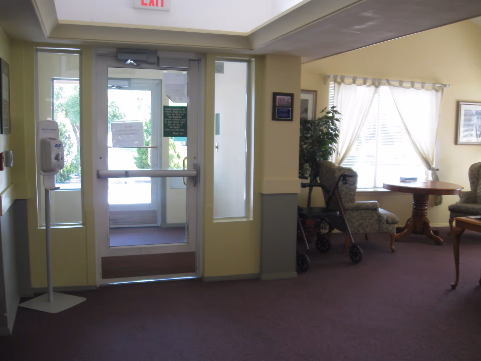 Interior view of a senior living facility entrance area with a glass door, exit sign above, a hand sanitizer dispenser on a stand to the left, and a sitting area with upholstered chairs, a wooden table, a walker, and a large window with sheer curtains on the right.
