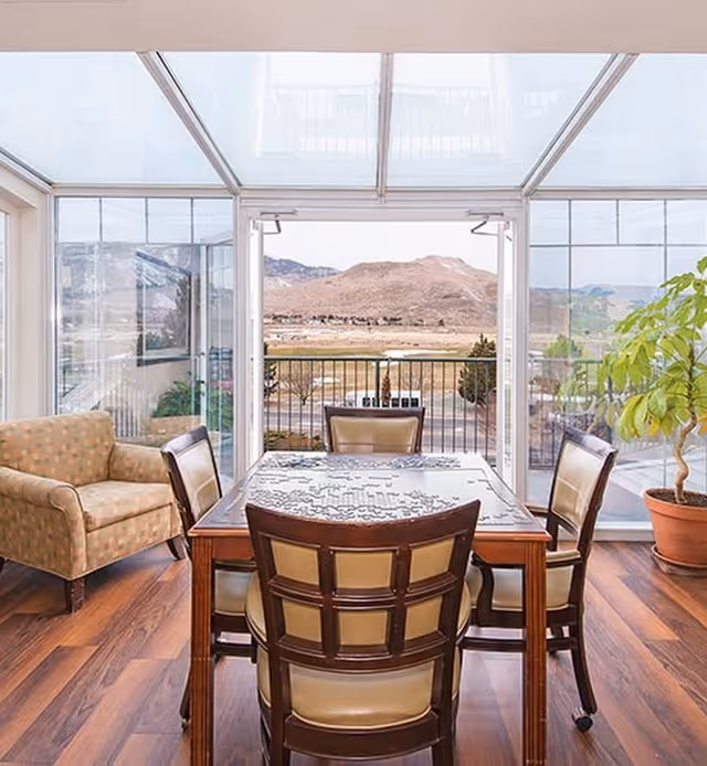 A bright room with large glass windows and a glass ceiling showing a scenic view of hills and trees outside. Inside, there is a wooden table with a partially completed jigsaw puzzle on it, surrounded by four cushioned chairs. To the left, there is a patterned armchair, and to the right, a potted plant on a wooden floor.