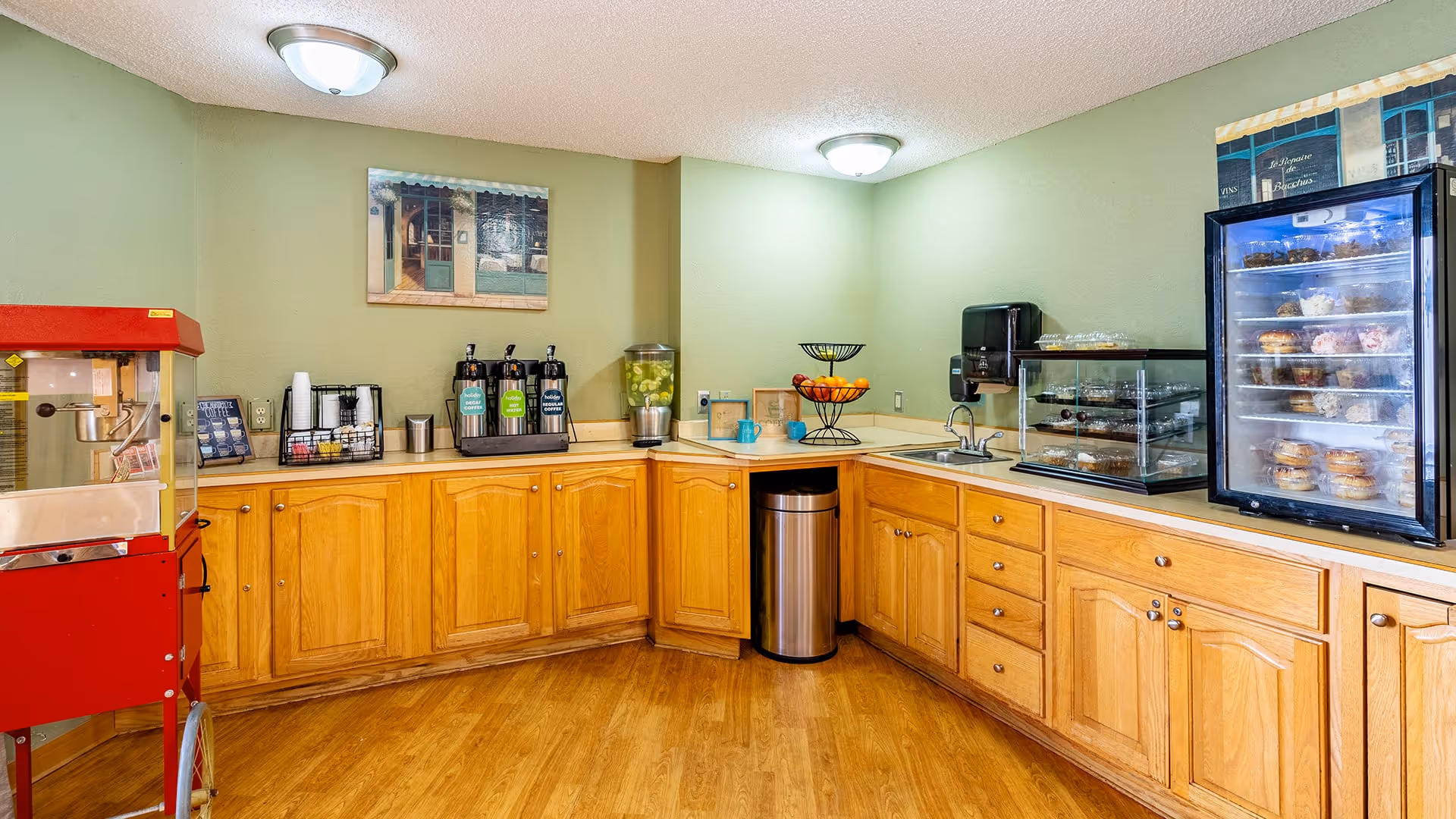 A kitchenette area with wooden cabinets and a light green wall. On the left side, there is a red popcorn machine. On the countertop, there are coffee dispensers, a water dispenser with lemon slices, a fruit basket, a small sink, a paper towel dispenser, and a display case with various pastries. The floor is wooden, and there are two ceiling lights illuminating the space.