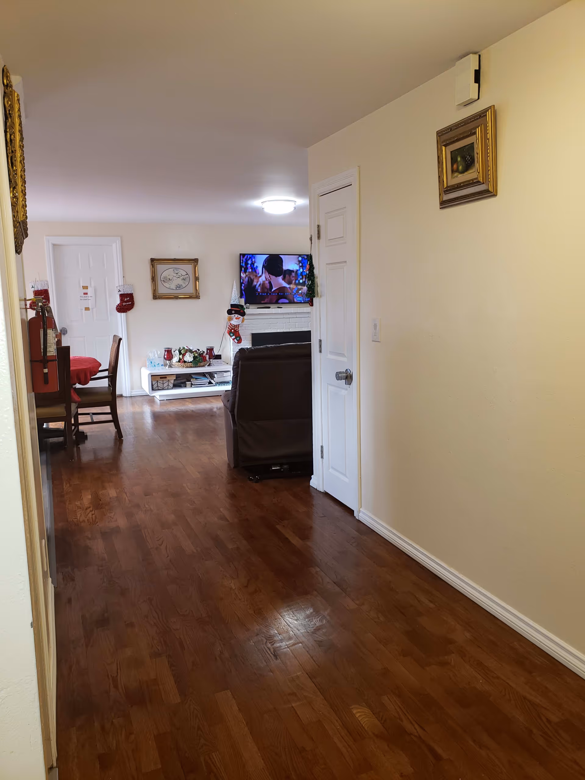 Interior view of a senior living facility hallway with wooden flooring leading to a living room area. The living room has a brown recliner chair facing a wall-mounted TV above a white fireplace decorated with Christmas stockings and a snowman figure. A dining table with chairs is visible to the left, covered with a red tablecloth. The walls are light-colored with framed artwork hanging.