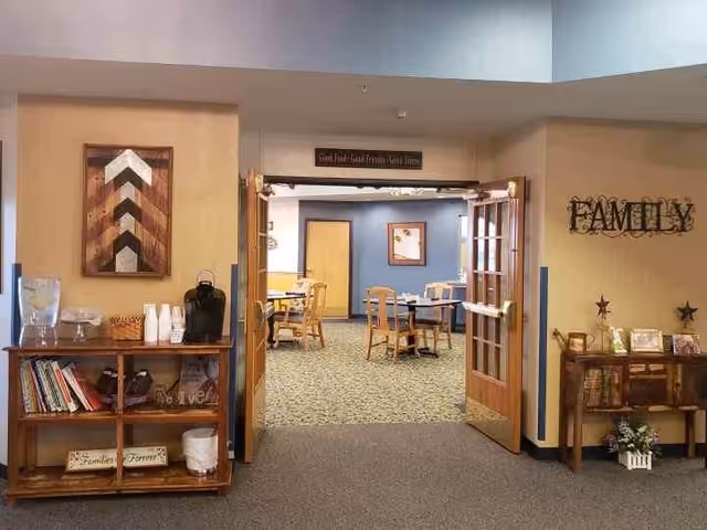 View through open double doors into a dining area with wooden tables and chairs. On the left side, there is a wooden shelf with books, cups, and a water dispenser. On the right side, there is a wooden cabinet with framed photos and decorative stars, with the word FAMILY mounted on the wall above it.