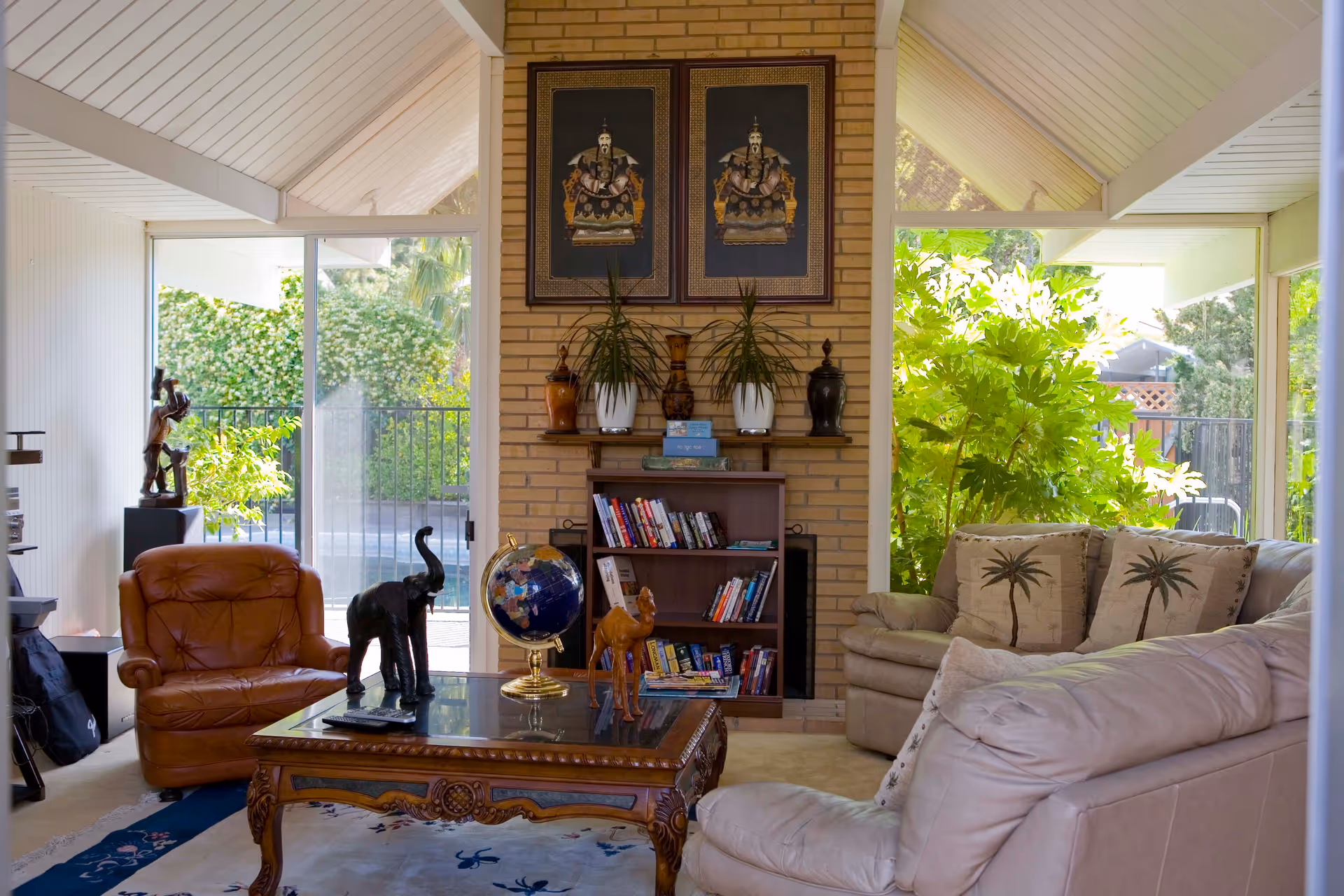 Bright living room with leather armchairs and sofas around a wooden coffee table, bookshelf and large windows showing greenery outside.