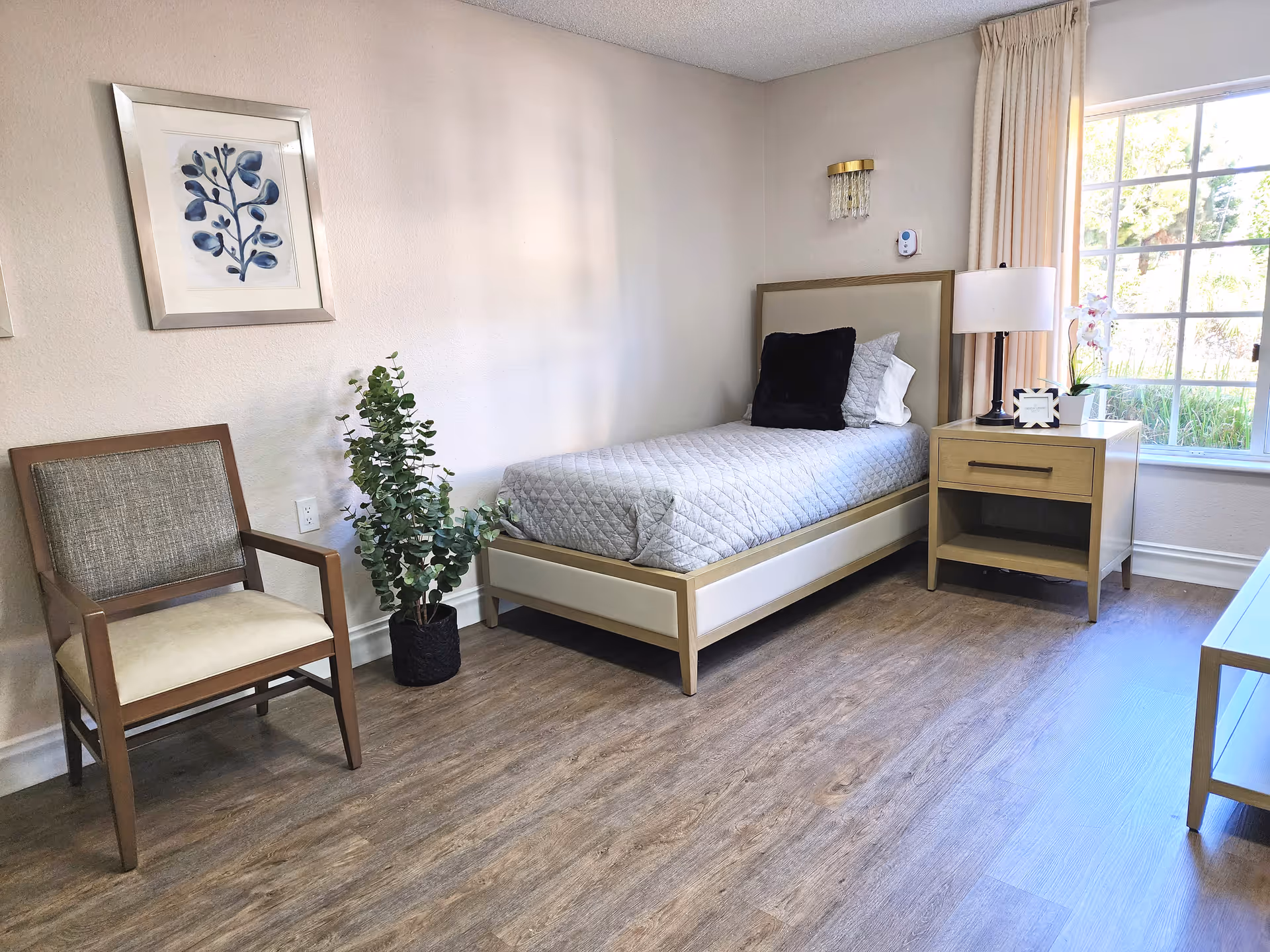 A bright and clean bedroom with a single bed covered in a gray quilt and pillows, a wooden nightstand with a lamp, a small plant, and a picture frame. There is a wooden chair with a cushioned seat and backrest, a potted plant on the floor, a framed botanical artwork on the wall, and a large window with beige curtains letting in natural light.
