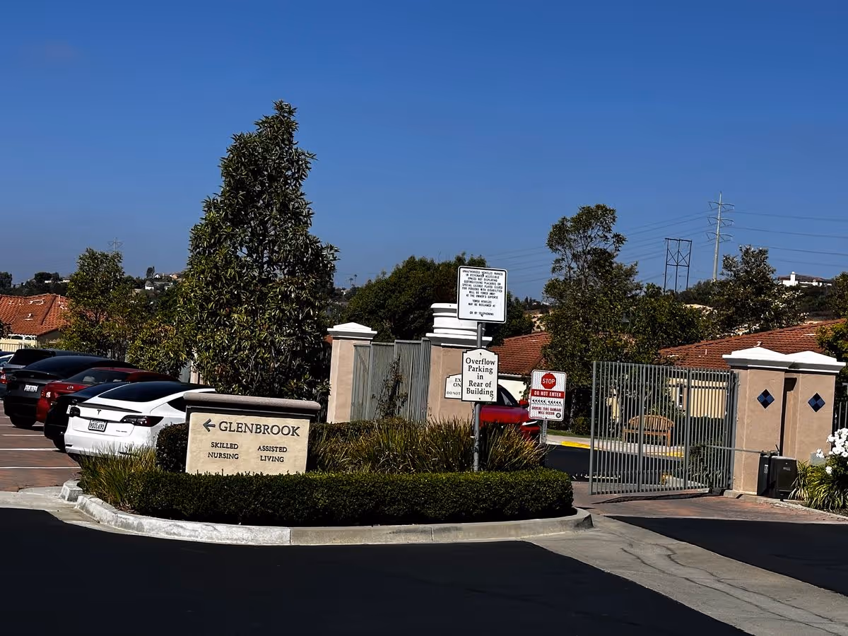 Entrance and gated parking area of GlenBrook Health Center with a stone sign reading 'GLENBROOK Skilled Nursing Assisted Living' and several parked cars.