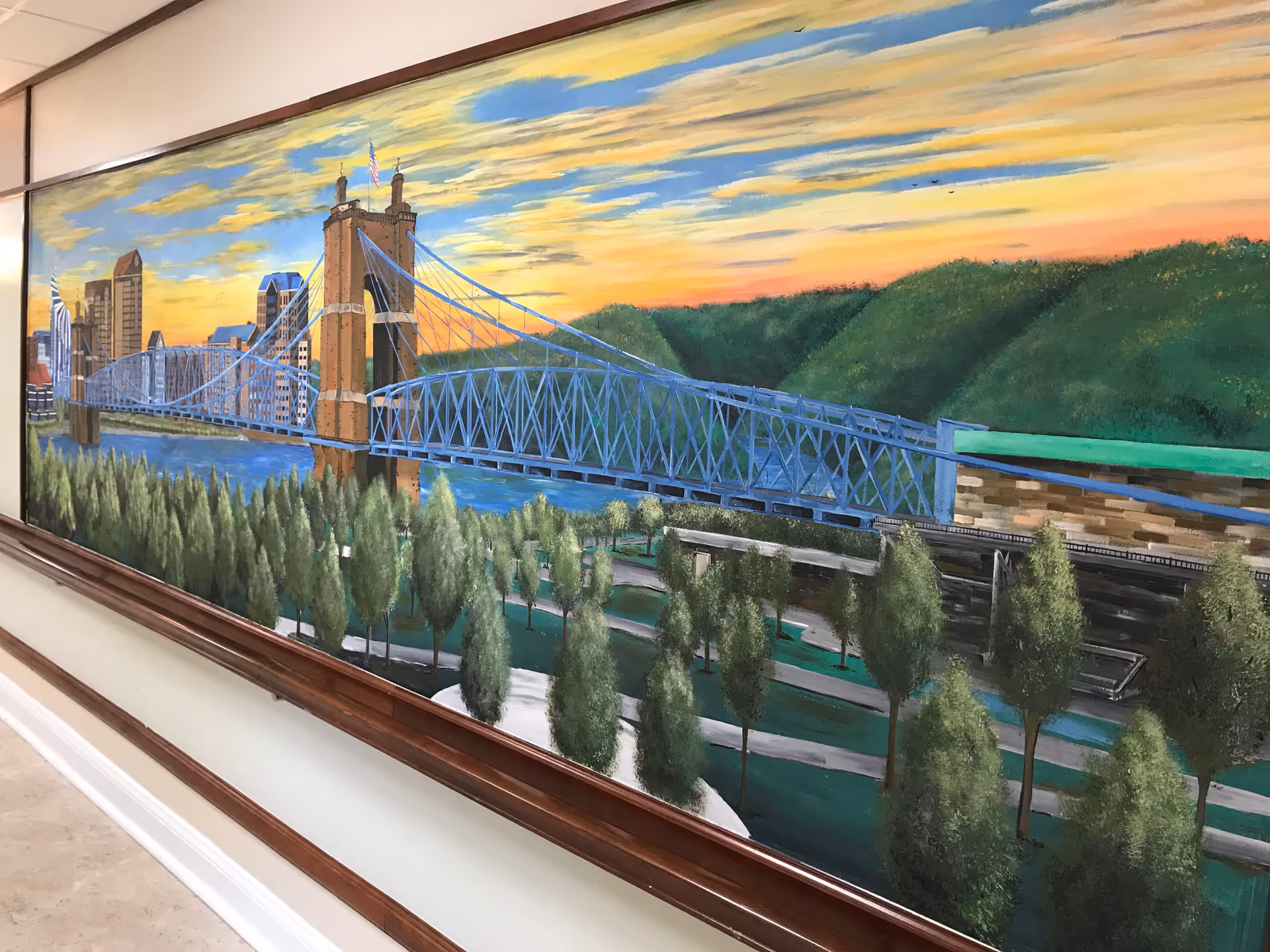 A large framed painting on a wall depicting a blue suspension bridge over a river with a city skyline in the background and green trees in the foreground under a colorful sunset sky.