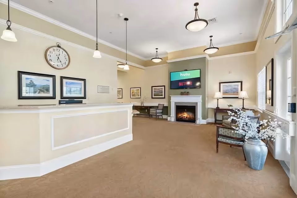 A bright and spacious reception area with a beige front desk on the left, a large clock and framed pictures on the wall behind it. The room features a cozy fireplace with a TV mounted above showing Hulu and Netflix logos. There are several chairs and tables arranged around the room, decorative lamps, and a large vase with white flowers near the entrance door on the right.