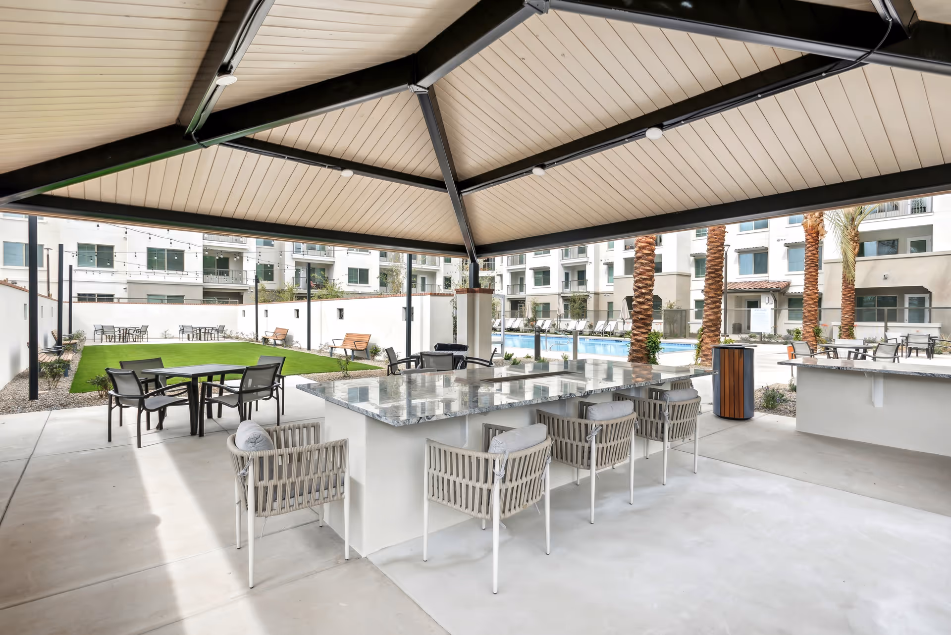 Outdoor covered seating area with a large stone countertop and cushioned chairs, overlooking a grassy lawn with benches and tables, palm trees, and a swimming pool surrounded by a multi-story residential building.
