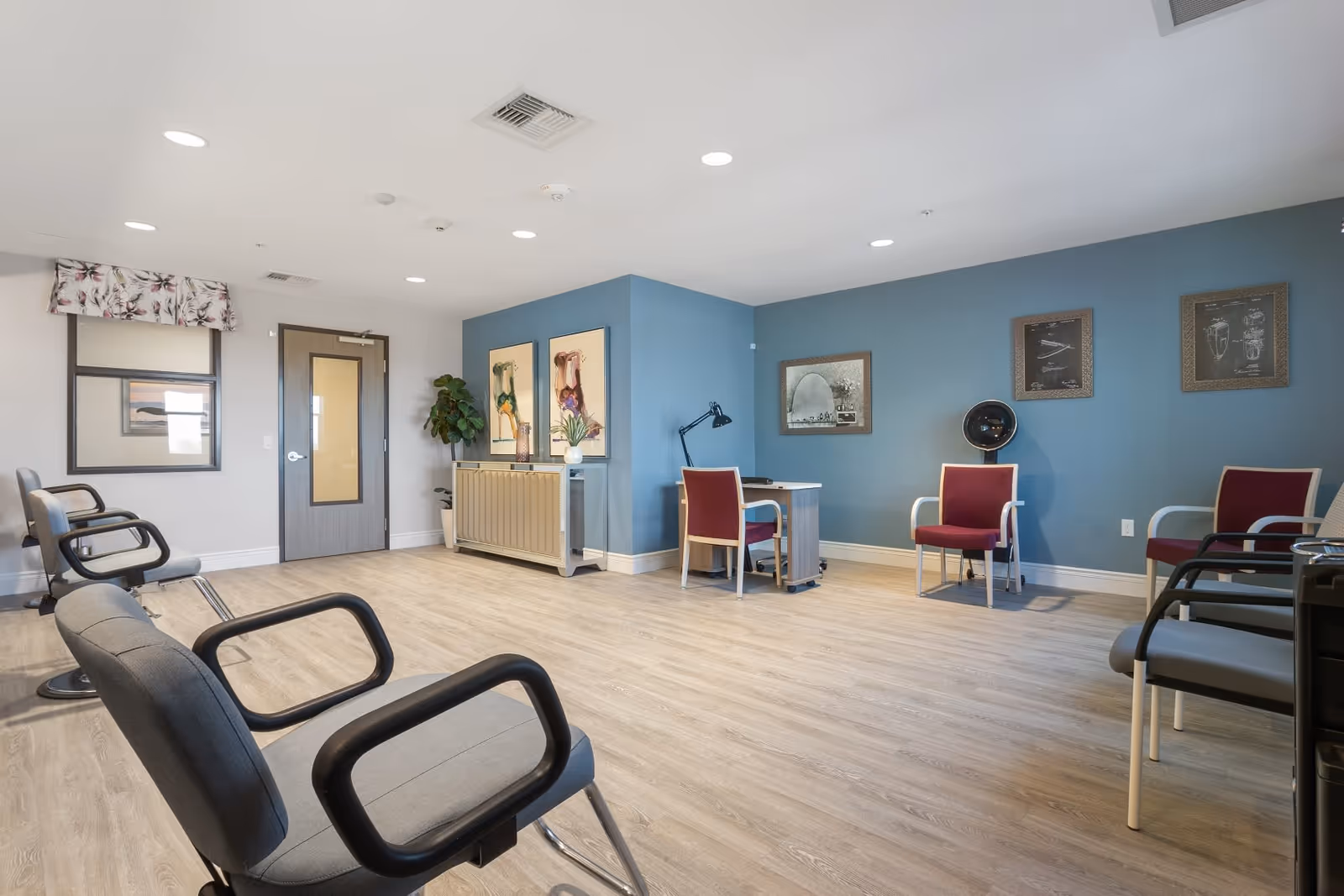 A spacious room with light wood flooring and a blue accent wall. The room contains several chairs arranged around the perimeter, including gray chairs with black armrests and maroon chairs with white armrests. There is a small desk with a black desk lamp and a chair, a cabinet with decorative items, framed artwork on the walls, and a potted plant near a door with a window. The ceiling has recessed lighting and air vents.