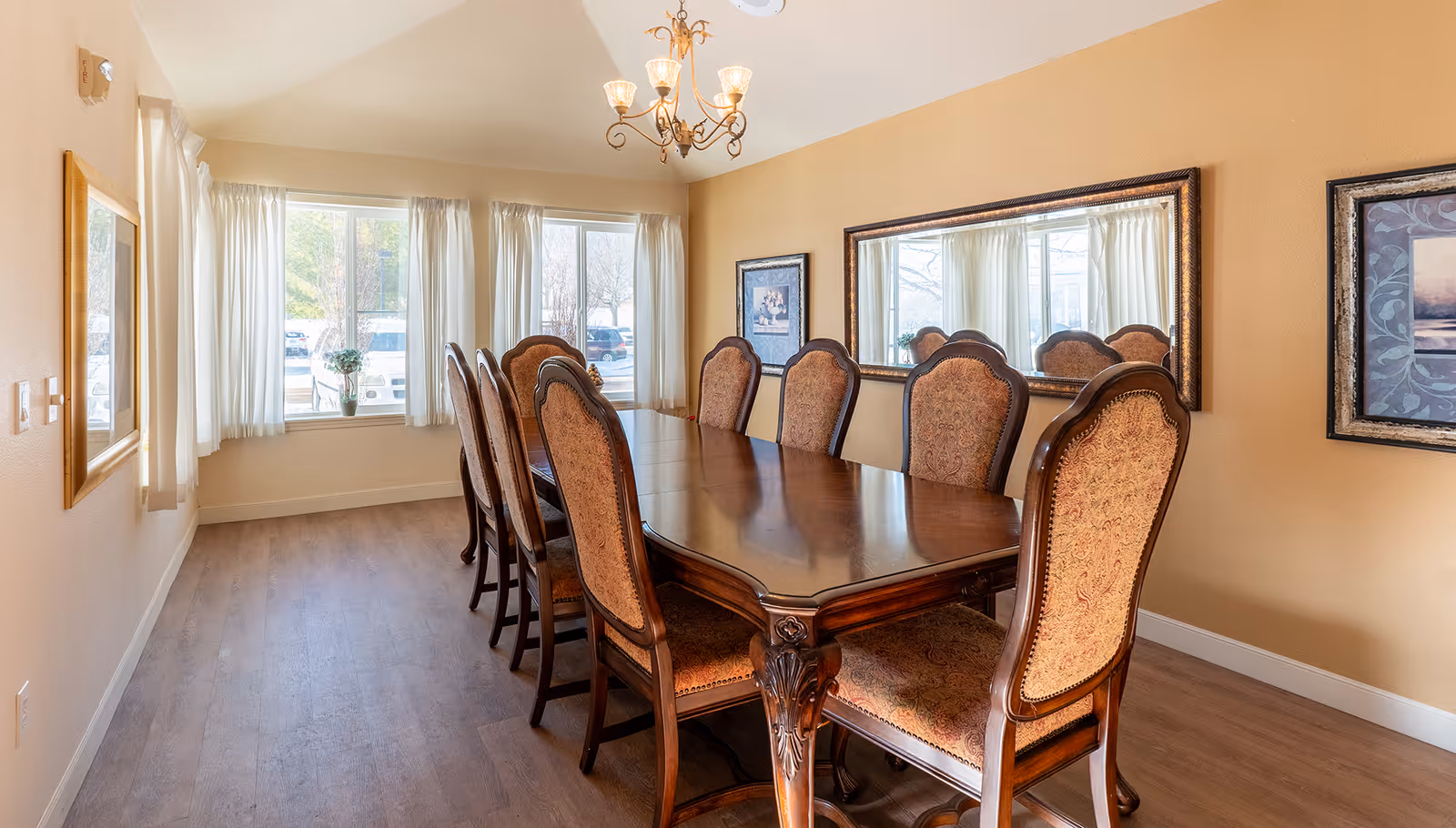 A well-lit dining room with a long wooden dining table surrounded by eight upholstered chairs. The room has large windows with white curtains allowing natural light to fill the space. There is a decorative chandelier hanging from the ceiling, a large mirror on one wall, and framed artwork on the walls. The floor is wooden, and the walls are painted a warm beige color.