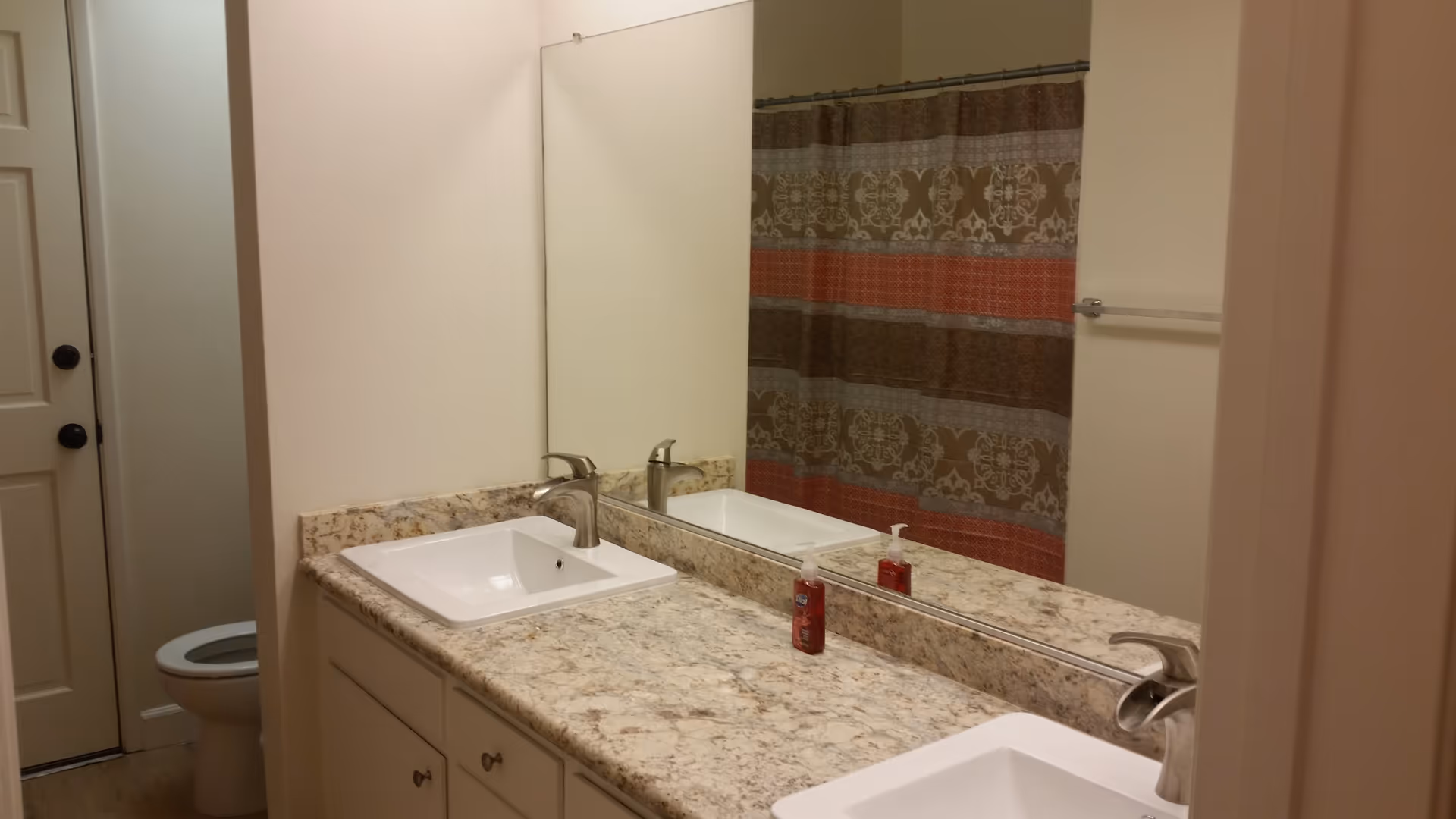 Bathroom with a granite countertop featuring two sinks and modern faucets, a large mirror above the sinks, a bottle of hand soap on the counter, a toilet partially visible behind a wall, and a shower curtain with a patterned design in shades of brown, red, and gray.