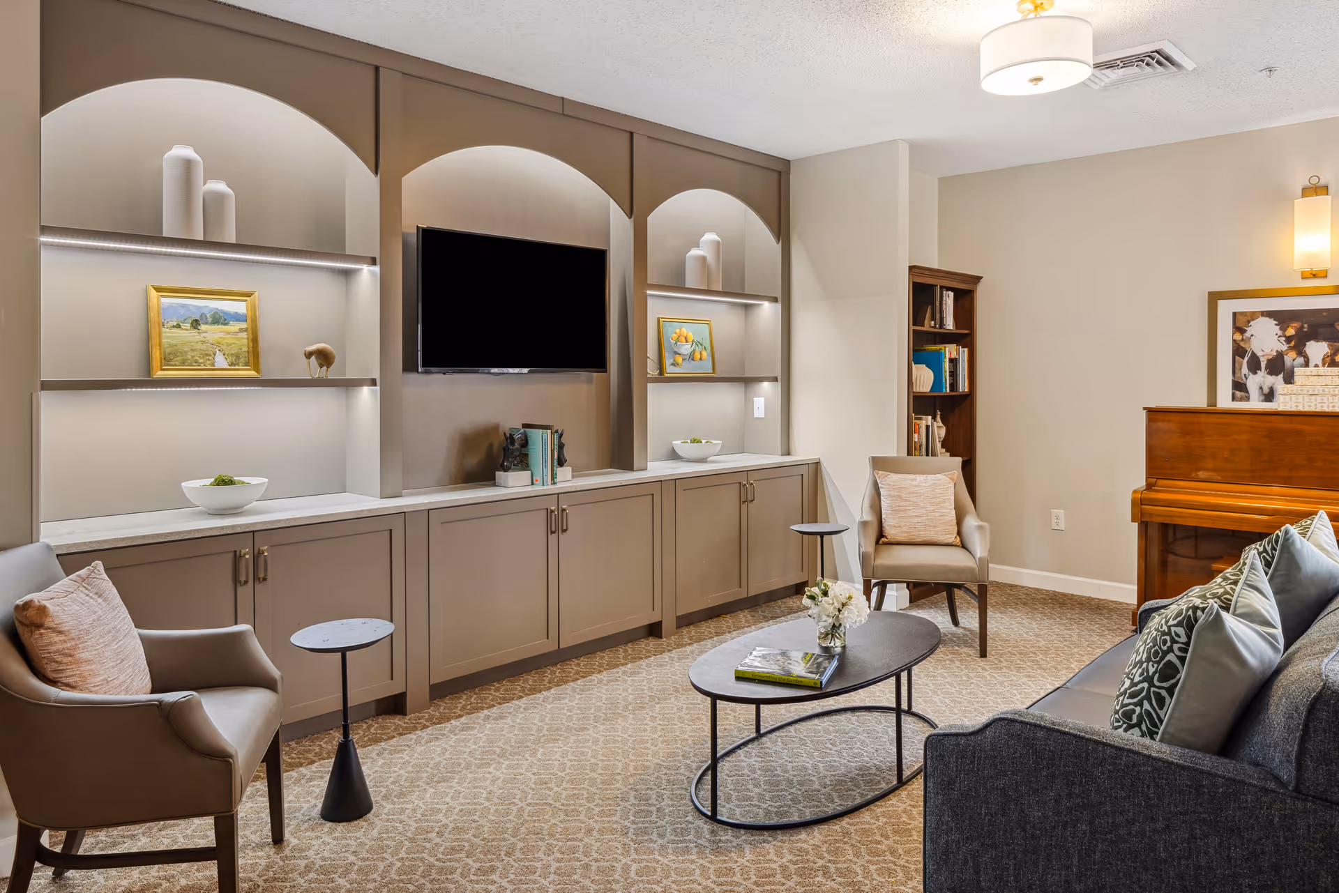 A cozy living room area with a gray sofa adorned with patterned pillows, two beige armchairs with cushions, a black oval coffee table with a flower vase and a book, built-in shelves with decorative items and framed paintings, a wall-mounted flat-screen TV, a wooden piano, and a bookshelf in the corner.