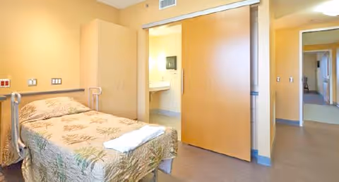 A single bedroom in a senior living facility with a bed covered in a floral bedspread and a folded white towel on top. The room has light yellow walls, a wooden wardrobe, and a sliding wooden door leading to a bathroom with a sink and mirror visible. The hallway outside the room is also visible.