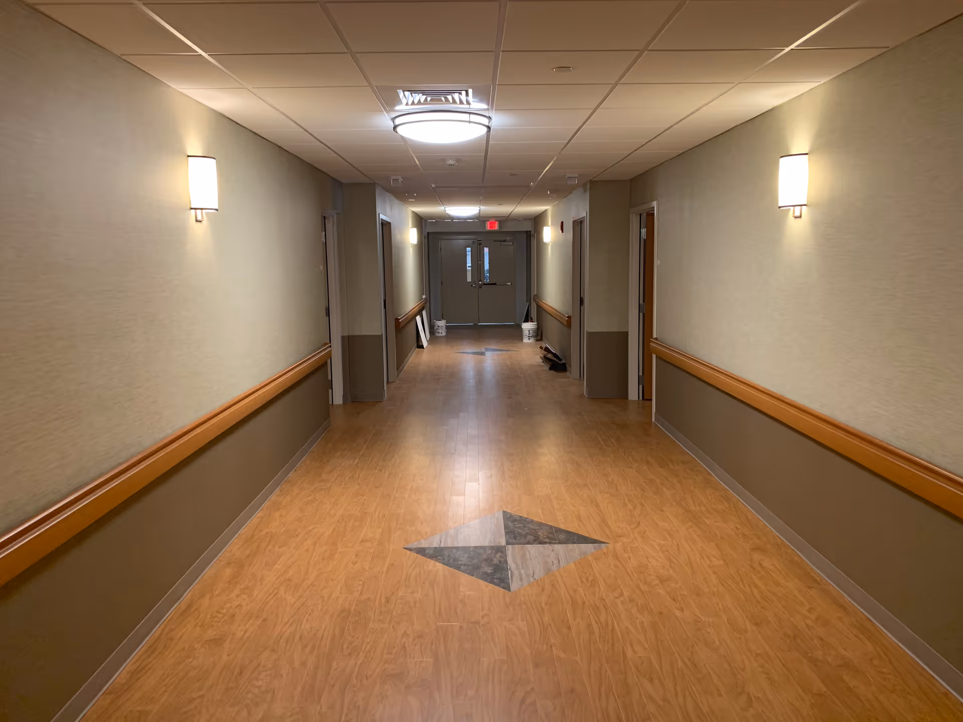 A well-lit indoor hallway with wood-patterned flooring and beige walls. The hallway has handrails on both sides and several doorways leading to rooms. Ceiling lights and wall sconces illuminate the space, and double doors are visible at the far end.