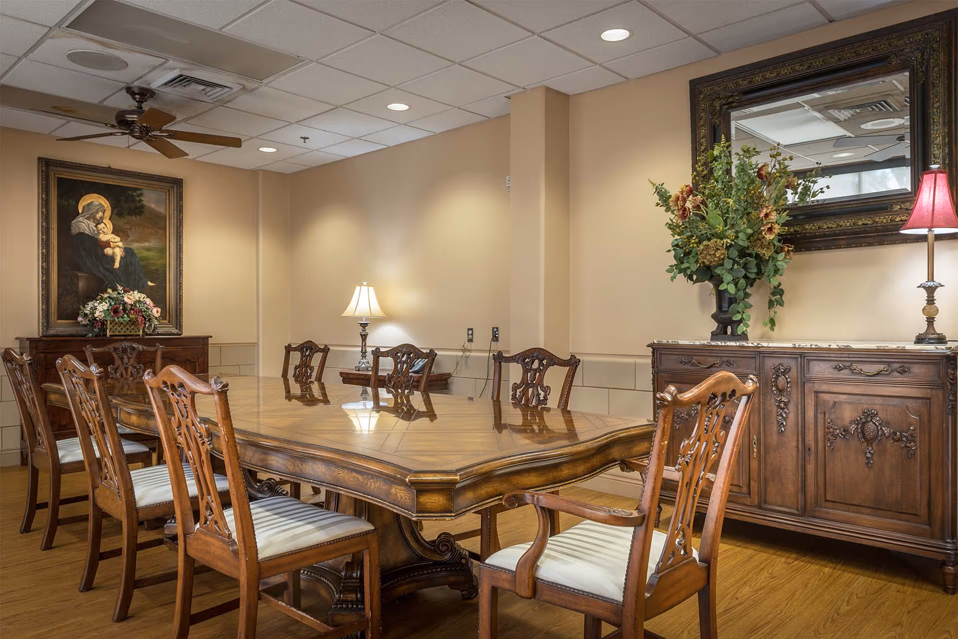 Ornate dining room with a large polished wooden table surrounded by carved chairs, sideboards, decorative lamps, a mirror, and framed artwork.