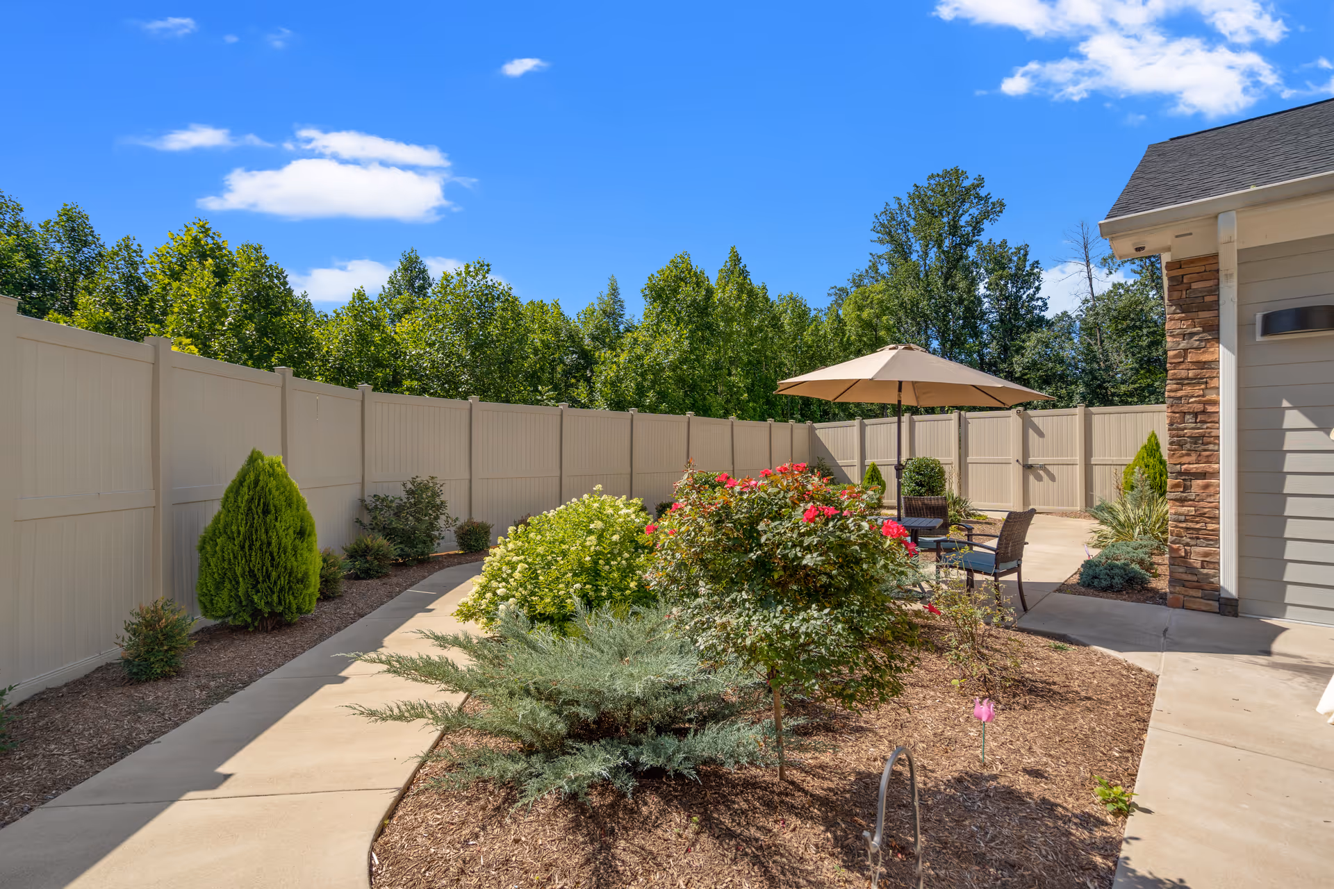 Sunlit fenced courtyard with landscaped planting beds, a paved walkway, and a patio seating area under an umbrella beside a building.