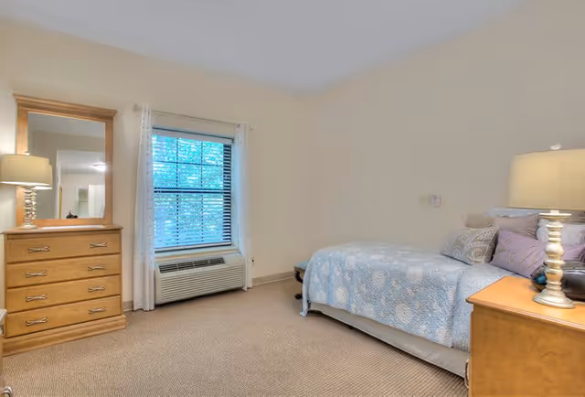 A cozy bedroom with a single bed covered in a light blue patterned quilt and several pillows. There is a wooden nightstand with a lamp on the right side of the bed. On the left side of the room, there is a wooden dresser with a mirror above it. A window with white curtains and blinds is centered on the wall, and below it is an air conditioning unit. The walls are painted a light beige color and the floor is carpeted.
