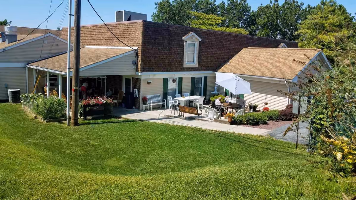 Exterior view of a single-story building with a brown shingled roof and beige siding. The building has a patio area with outdoor furniture including tables, chairs, benches, and a large white umbrella. There is a well-maintained green lawn in the foreground and some trees and shrubs surrounding the building.
