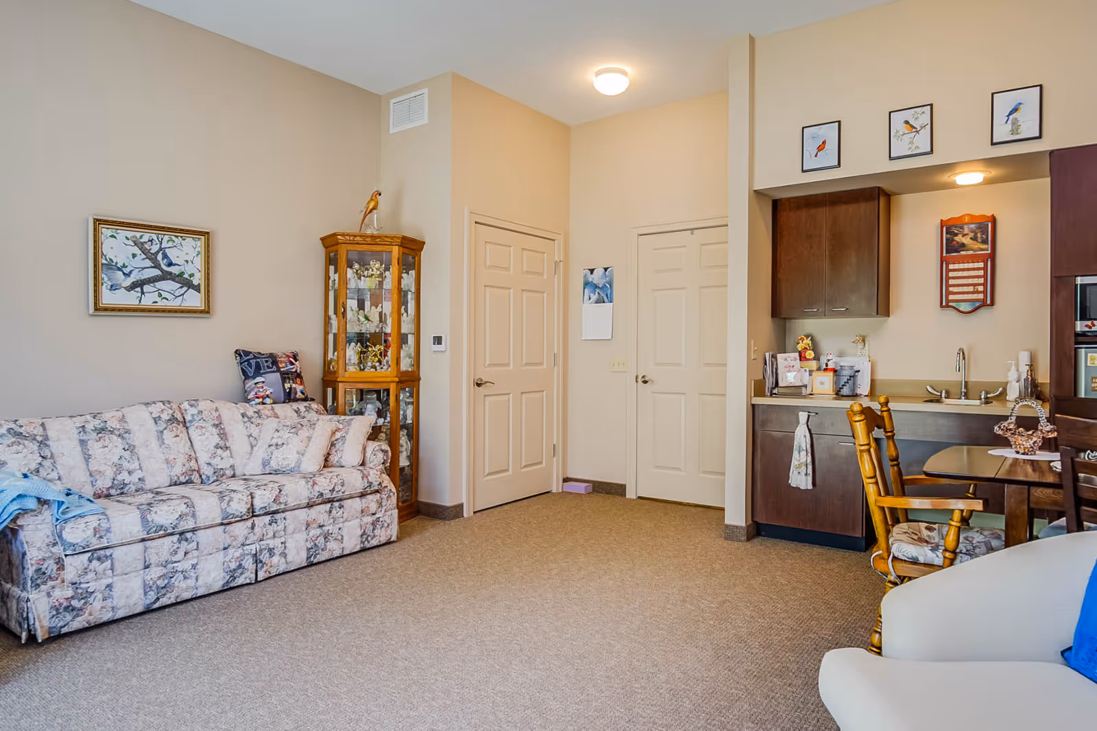 Bright assisted-living living room with a floral sofa, glass display cabinet, two doors, and a small kitchenette with dining chairs.