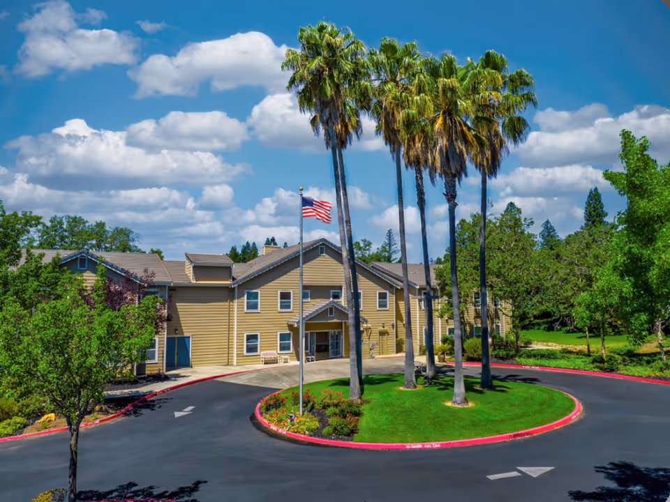 Front exterior of a yellow senior living building with tall palm trees, an American flag, and a circular driveway.