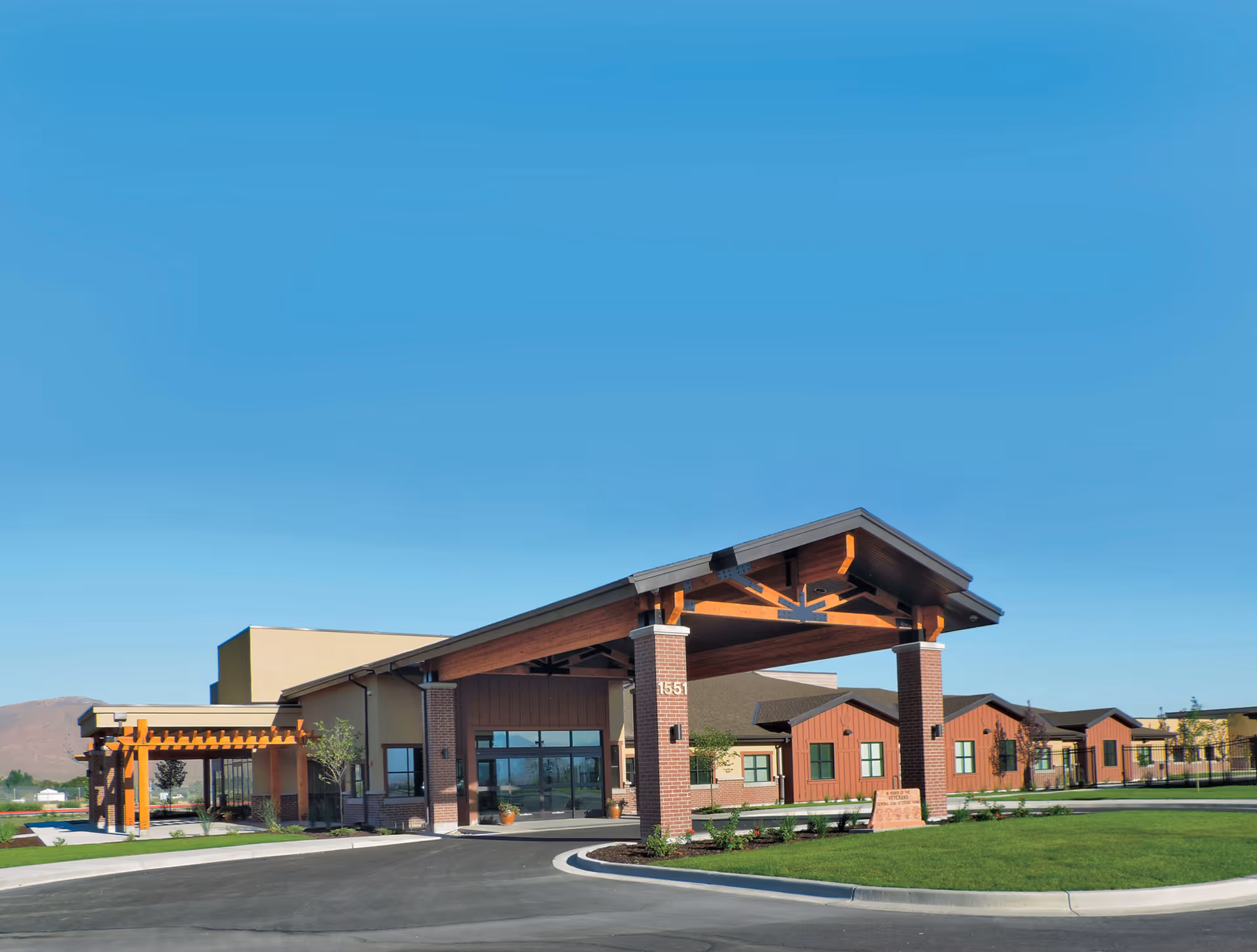 Exterior view of the Central Utah Veterans Home building with a large covered entrance supported by brick pillars, a well-maintained lawn, and clear blue sky in the background.