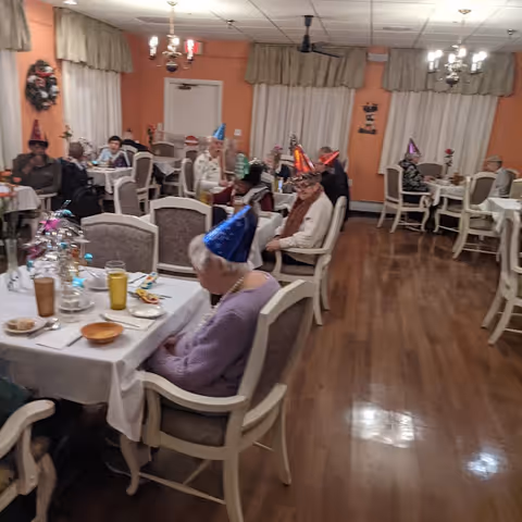 Dining room with elderly residents wearing party hats seated at tables set for a meal.