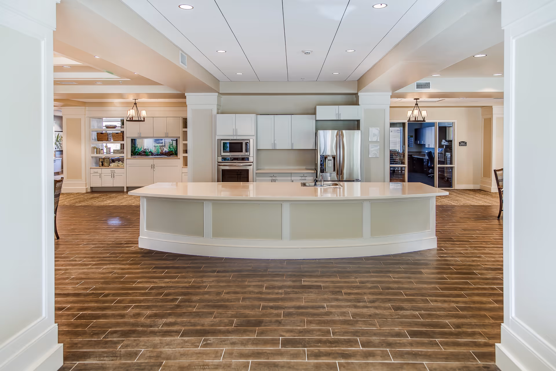 A spacious and modern kitchen area in a senior living facility with a large curved island countertop, white cabinetry, stainless steel refrigerator, built-in microwave, and oven. The floor has wood-like tiles and the ceiling has recessed lighting. There are open shelves and an aquarium on the left side, and an office area visible through a doorway on the right.