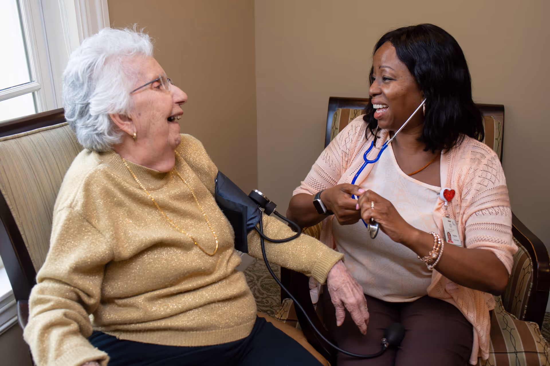 An elderly woman sitting in a chair with a blood pressure cuff on her arm, smiling and interacting with a healthcare worker who is using a stethoscope and smiling back at her in a warmly lit room.