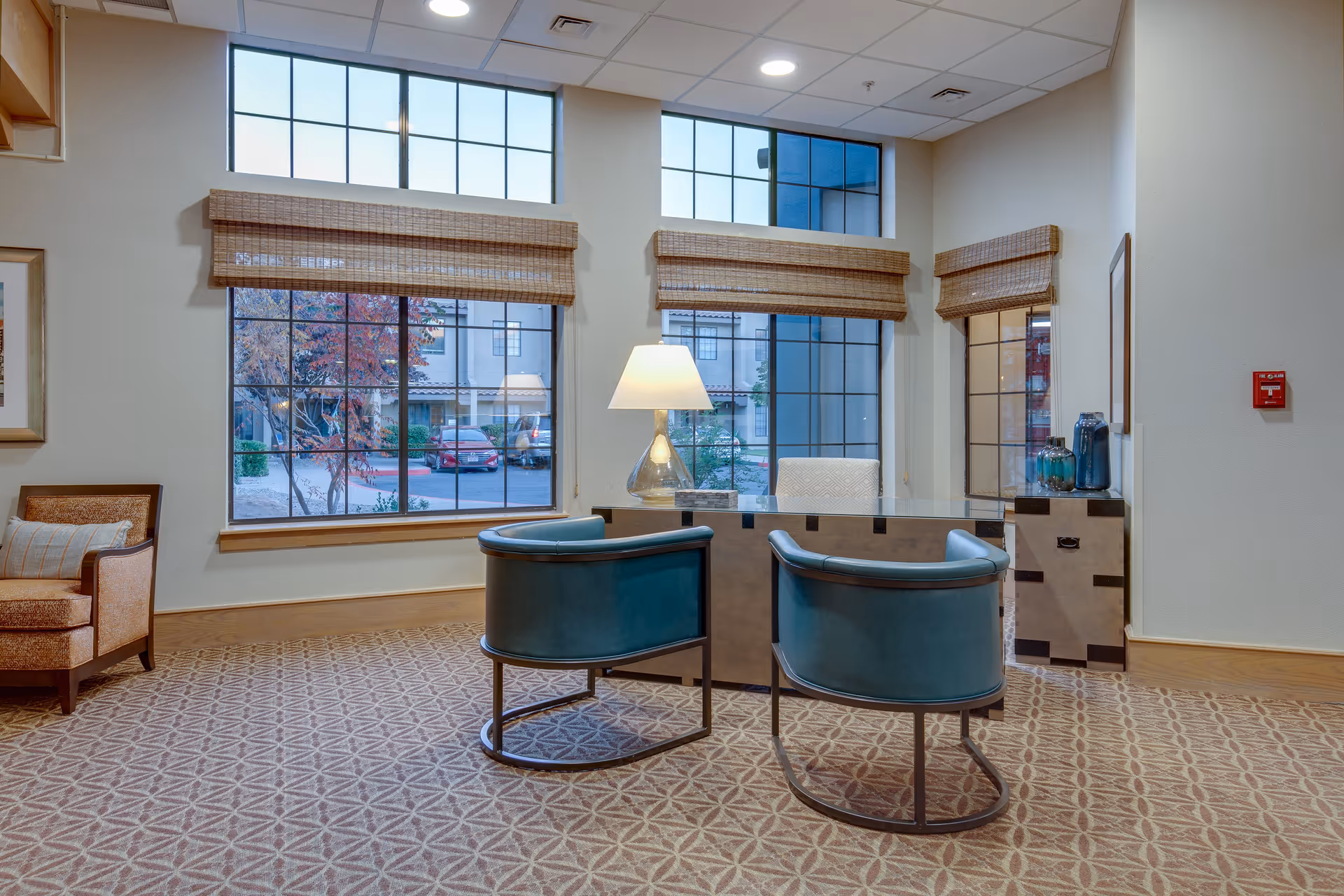 A bright interior room with large windows showing an outdoor view. The room features a desk with a lamp and two teal chairs in front of it. There is a beige armchair with a pillow to the left and a small cabinet with decorative items to the right. The floor is carpeted with a patterned design.