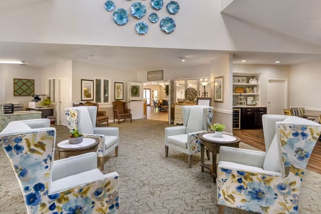 Sunlit communal lounge with floral-upholstered armchairs arranged around small tables beneath decorative wall art.