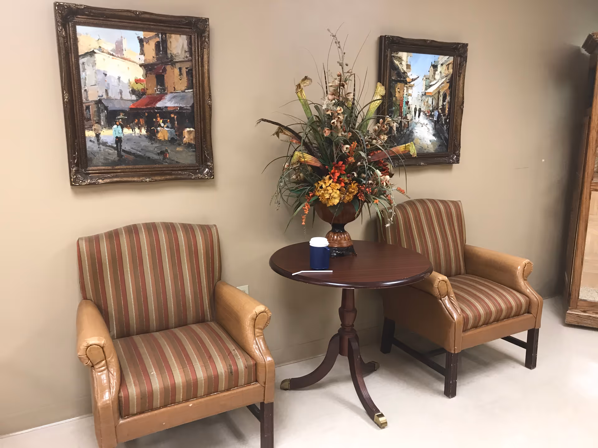 Two striped armchairs flank a small round table holding a floral arrangement and a coffee cup beneath two framed paintings in an indoor sitting area.