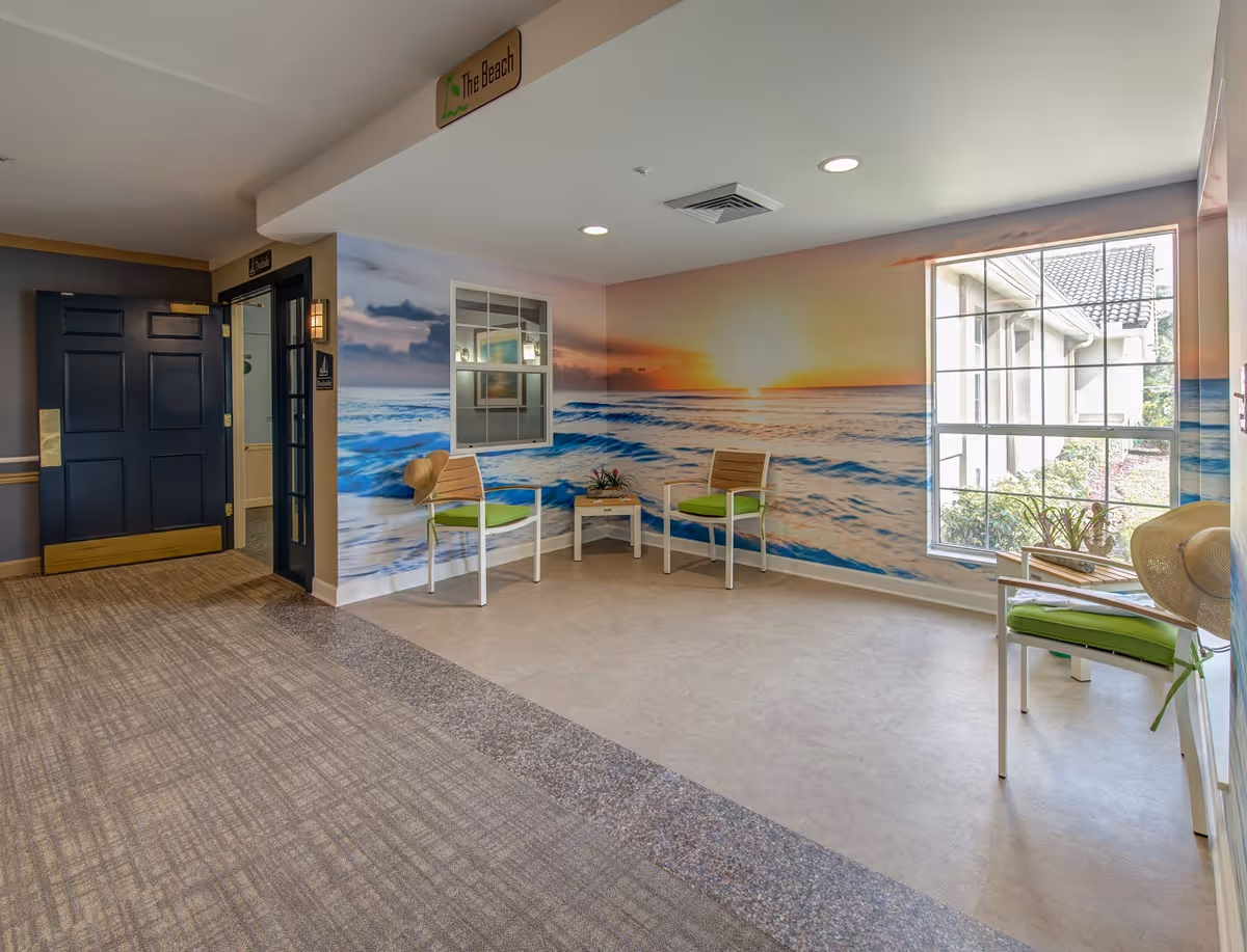 A bright indoor seating area with a large wall mural of a sunset over the ocean. The space has three chairs with green cushions and a small table with a plant. There is a window showing the outside of the building and a door leading to another room. A sign above reads 'The Beach'.