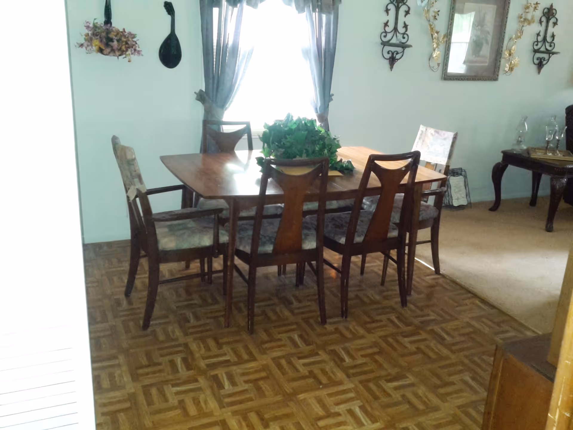 A dining room with a wooden dining table and six chairs, some with armrests and some without. The table has a green leafy centerpiece. The room has parquet flooring and light-colored walls with decorative wall hangings and framed artwork. A window with sheer curtains allows natural light into the room.