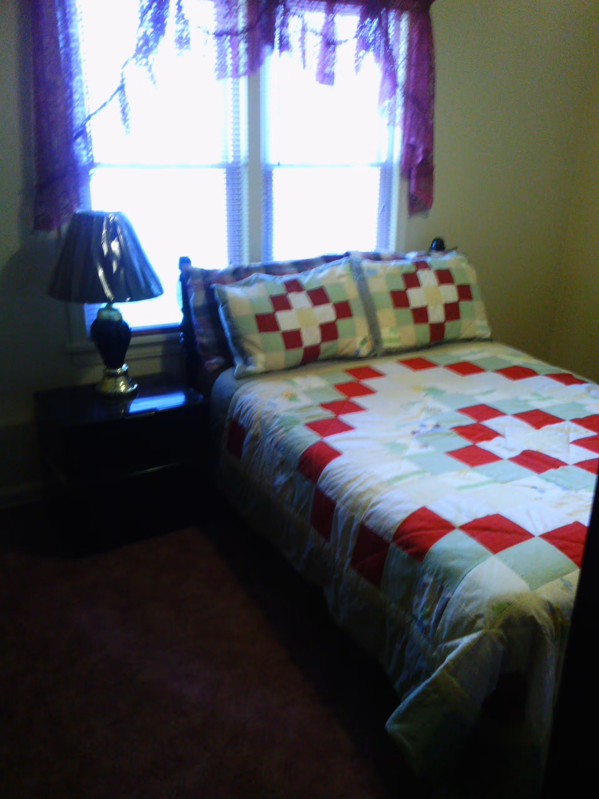 A small bedroom with a bed covered in a red, white, and light blue patchwork quilt and matching pillows. There is a dark wooden nightstand next to the bed with a black lamp on it. A window with purple lace curtains is behind the bed.