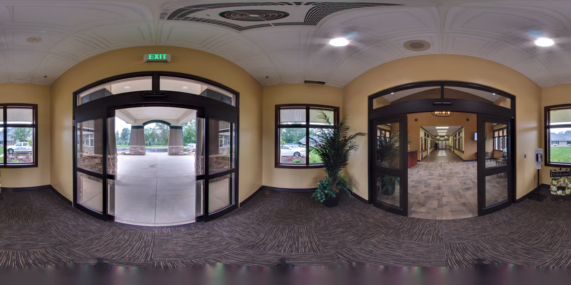 Entrance area of Miller's Merry Manor showing double glass doors leading outside to a covered driveway with parked cars visible. Inside, there is a hallway with carpeted floor and beige walls, a potted plant near a window, and another set of glass doors leading further into the building.