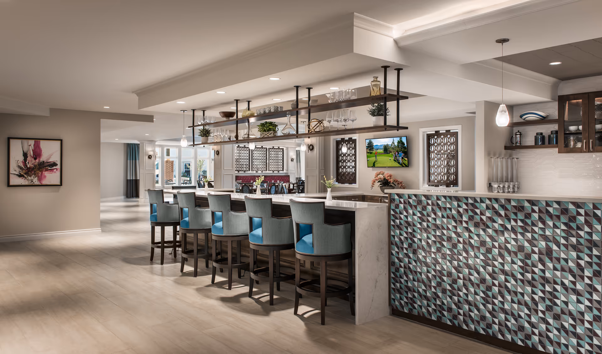 Open communal dining and bar area with a long marble-topped counter, blue-cushioned bar stools, hanging shelves with glassware, and seating in the background.
