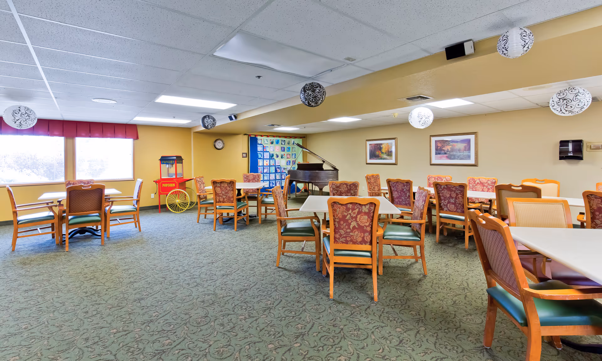 Spacious senior living common room with multiple tables and patterned chairs, a grand piano, popcorn cart, and hanging decorative lights.