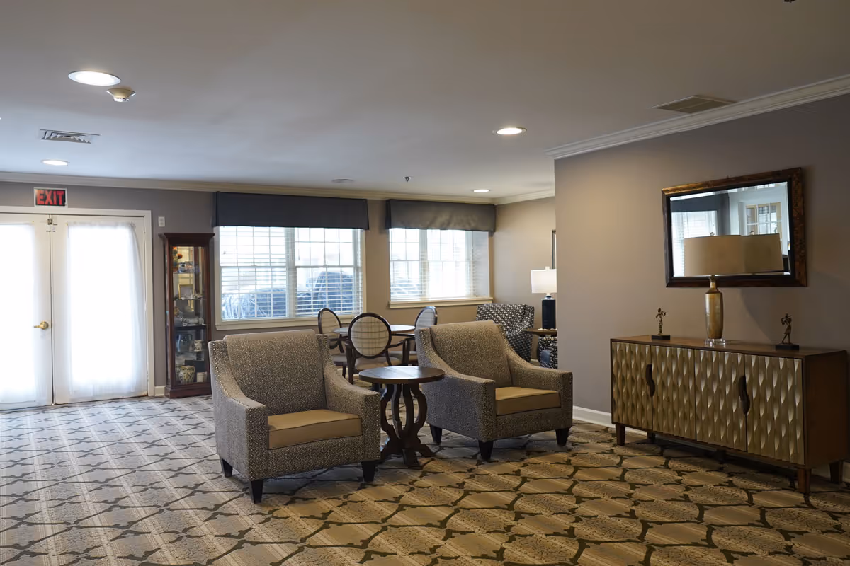 A spacious and well-lit common area in a senior living facility featuring two upholstered armchairs with a small round wooden table between them, a patterned carpet, a sideboard with a decorative lamp and small sculptures, a large mirror on the wall, and a seating area with a table and chairs near windows with blinds.
