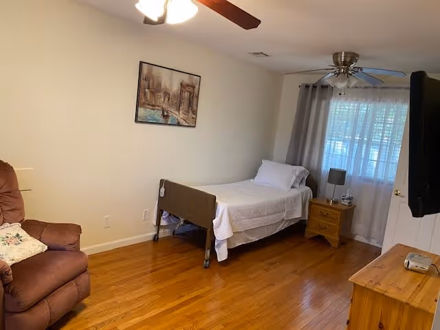 A simple bedroom with a single bed with safety rails, a recliner, wooden nightstand and dresser, hardwood floors, and a window with curtains.