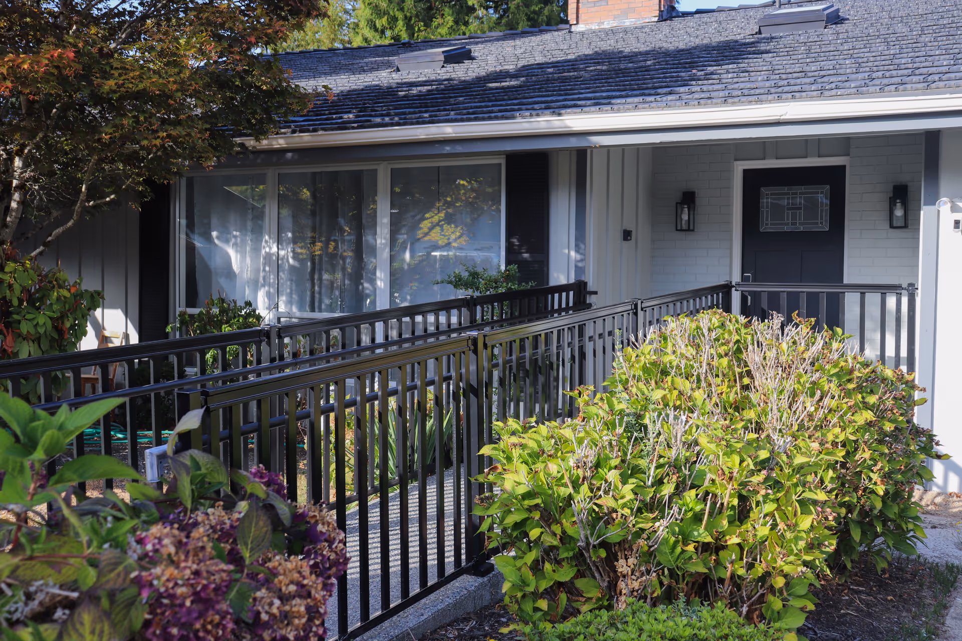 Front exterior view of a single-story building with a black door, large windows, and a black metal railing along a ramp leading to the entrance. There are green bushes and plants in the foreground and a tree to the left side of the image.
