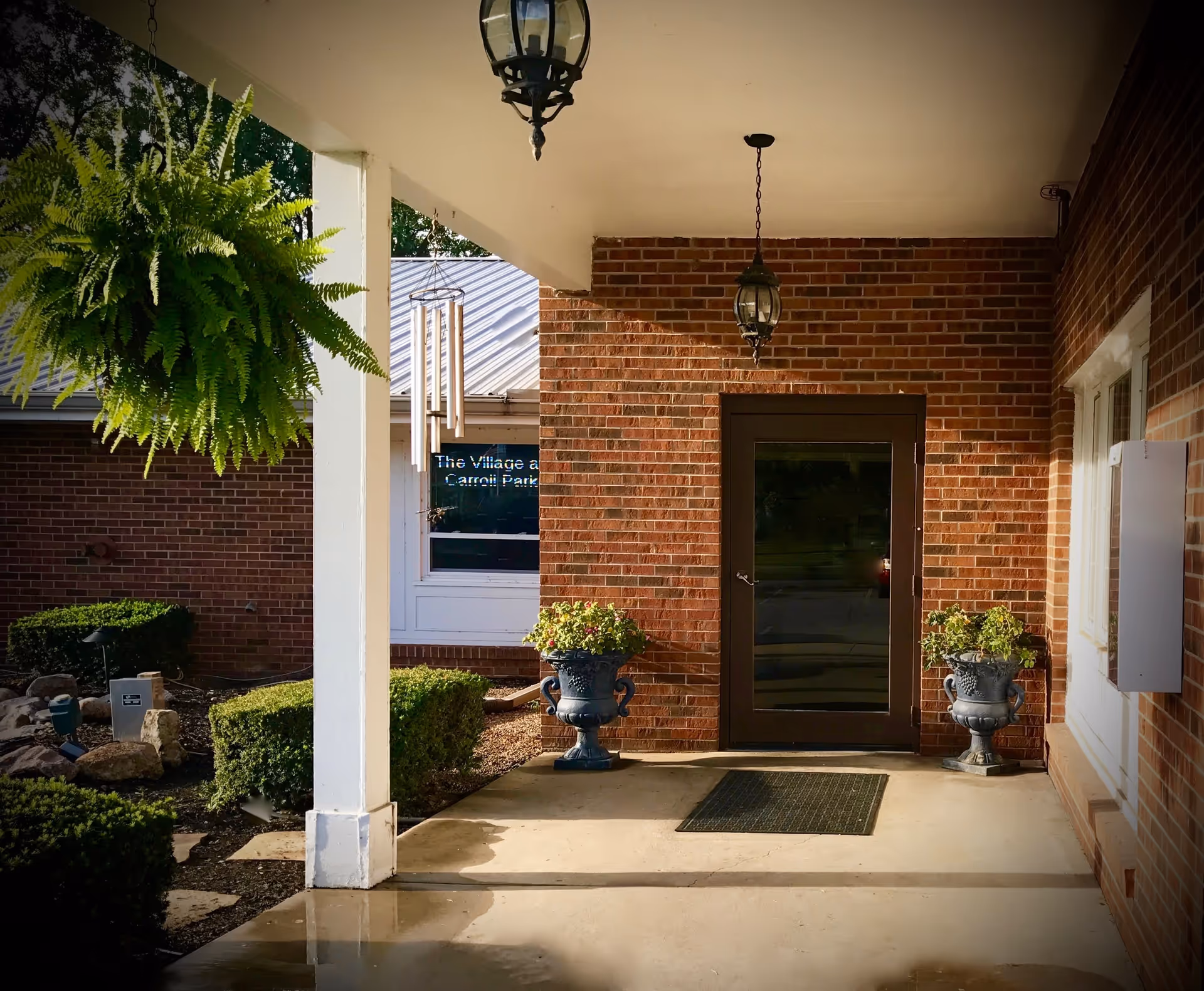 Covered entrance area of a brick building with a glass door, two potted plants on either side of the door, hanging lantern lights, a hanging fern plant, and a wind chime. There are trimmed bushes and a small garden area visible to the left.