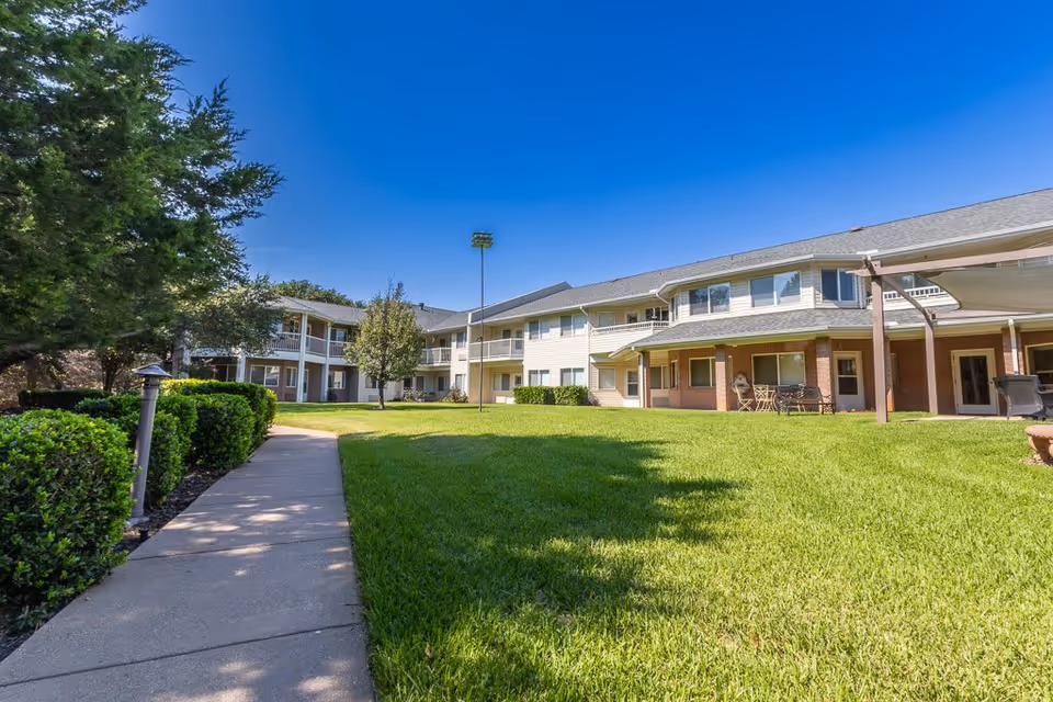 Outdoor view of Asher Point Independent Living of Arlington showing a well-maintained lawn, a paved walkway, bushes, trees, and a two-story building with balconies and a covered patio area under a clear blue sky.