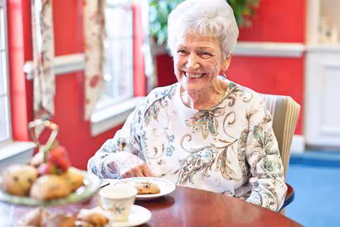 An elderly woman with white hair is sitting at a dining table in a room with red walls and large windows. She is smiling and wearing a floral patterned top. On the table in front of her are a plate with a pastry, a teacup, and a tiered tray with more pastries.