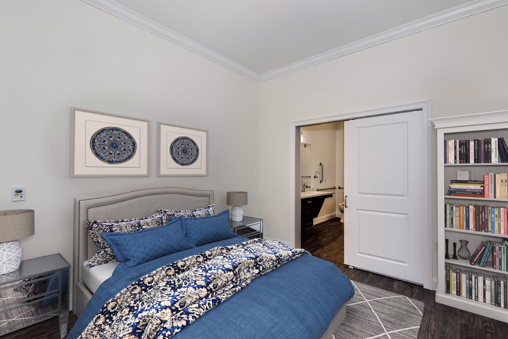 A bedroom with a neatly made bed featuring blue and floral bedding. The bed has a gray upholstered headboard and is flanked by two mirrored nightstands with matching lamps. On the wall above the bed are two framed decorative artworks. To the right, there is a white sliding door partially open, revealing a bathroom with a sink and mirror. A white bookshelf filled with books is positioned next to the door. The room has light-colored walls and dark wood flooring with a gray patterned rug.