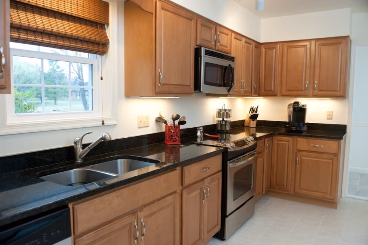 A clean kitchen with wooden cabinets, black granite countertops, a double sink under a window with bamboo blinds, a stainless steel stove with an oven, a microwave above it, a coffee maker, a knife block, and various kitchen utensils on the counter.