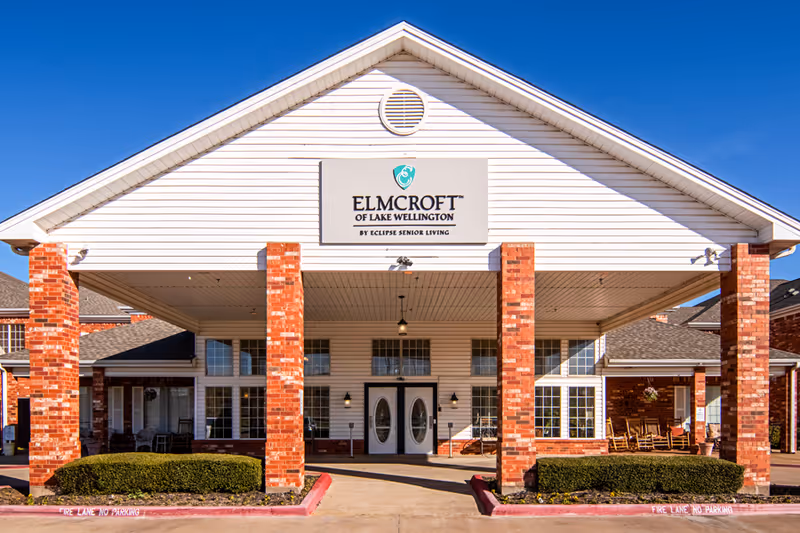 Front exterior view of a senior living facility named Elmcroft of Lake Wellington, featuring a white gabled entrance with red brick columns and large windows under a clear blue sky.