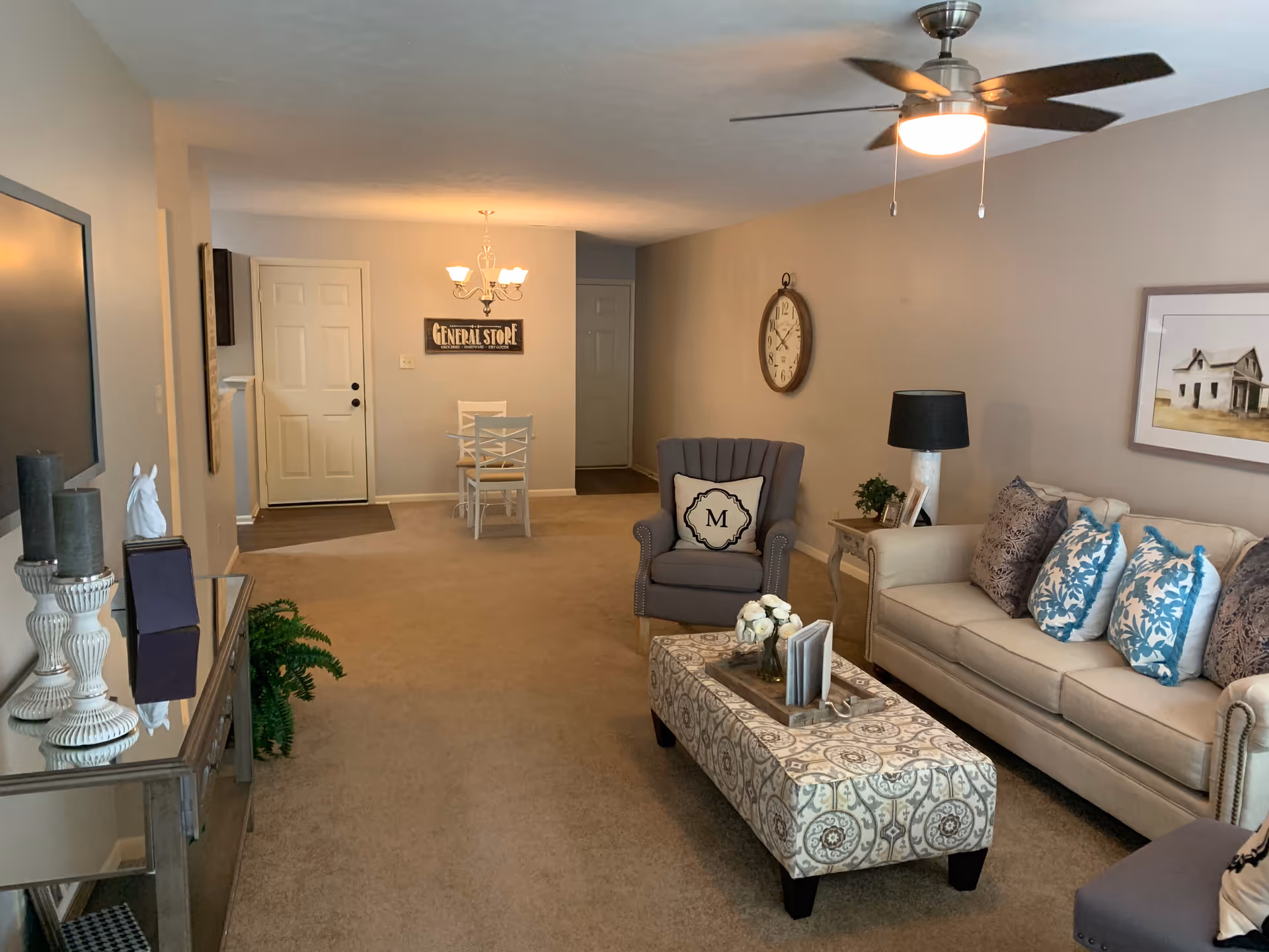 A cozy living room with a beige sofa adorned with blue and patterned pillows, a matching armchair with a pillow marked 'M', a patterned ottoman with a tray holding flowers and books, a side table with a lamp and framed photos, a wall clock, and a framed picture of a house. In the background, there is a small dining area with a white table and chairs under a chandelier, and a door with a 'General Store' sign above it. The room has beige carpet and neutral-colored walls.
