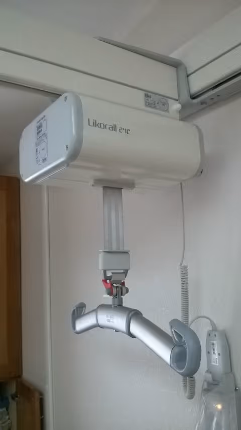 A ceiling-mounted patient lift device labeled 'Likorall 242' with a horizontal metal bar and curved handles, installed in a room with white walls and a wooden cabinet partially visible on the left.