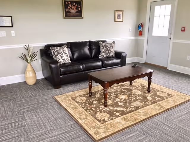 A living room area with a black leather sofa adorned with two patterned cushions, a wooden coffee table on a decorative area rug, a vase with tall plants on the left, and a door with a window on the right. The walls are light-colored with white trim, and there are two framed pictures hanging above the sofa.