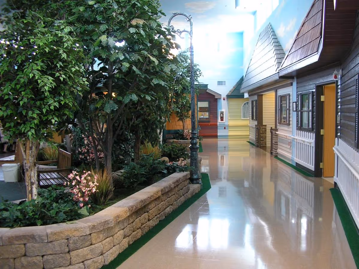 Indoor hallway designed to resemble an outdoor street scene with small house facades on the right, a garden area with trees and flowers on the left, and a street lamp in the center under a ceiling painted with blue sky and clouds.